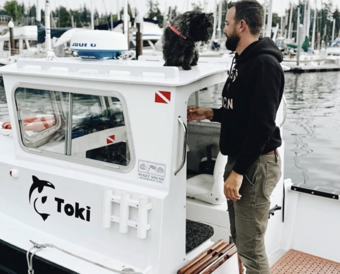 Bearded man standing on a small white boat at a marina, a small black dog with a red collar sits on the roof beside him.