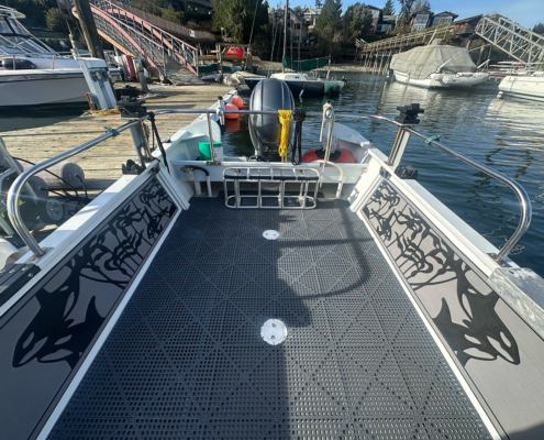 Deck of a small motorboat docked at a marina, showing an outboard motor at the stern and metal grid flooring with decorative side panels, nearby boats and docks in the background.