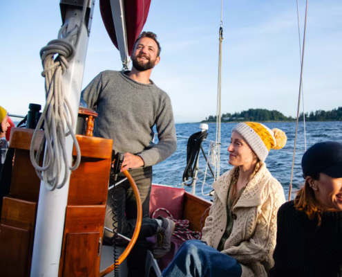 Smiling man at the helm steering a sailboat with two women seated nearby, blue ocean and distant island in the background.