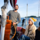 Smiling man at the helm steering a sailboat with two women seated nearby, blue ocean and distant island in the background.