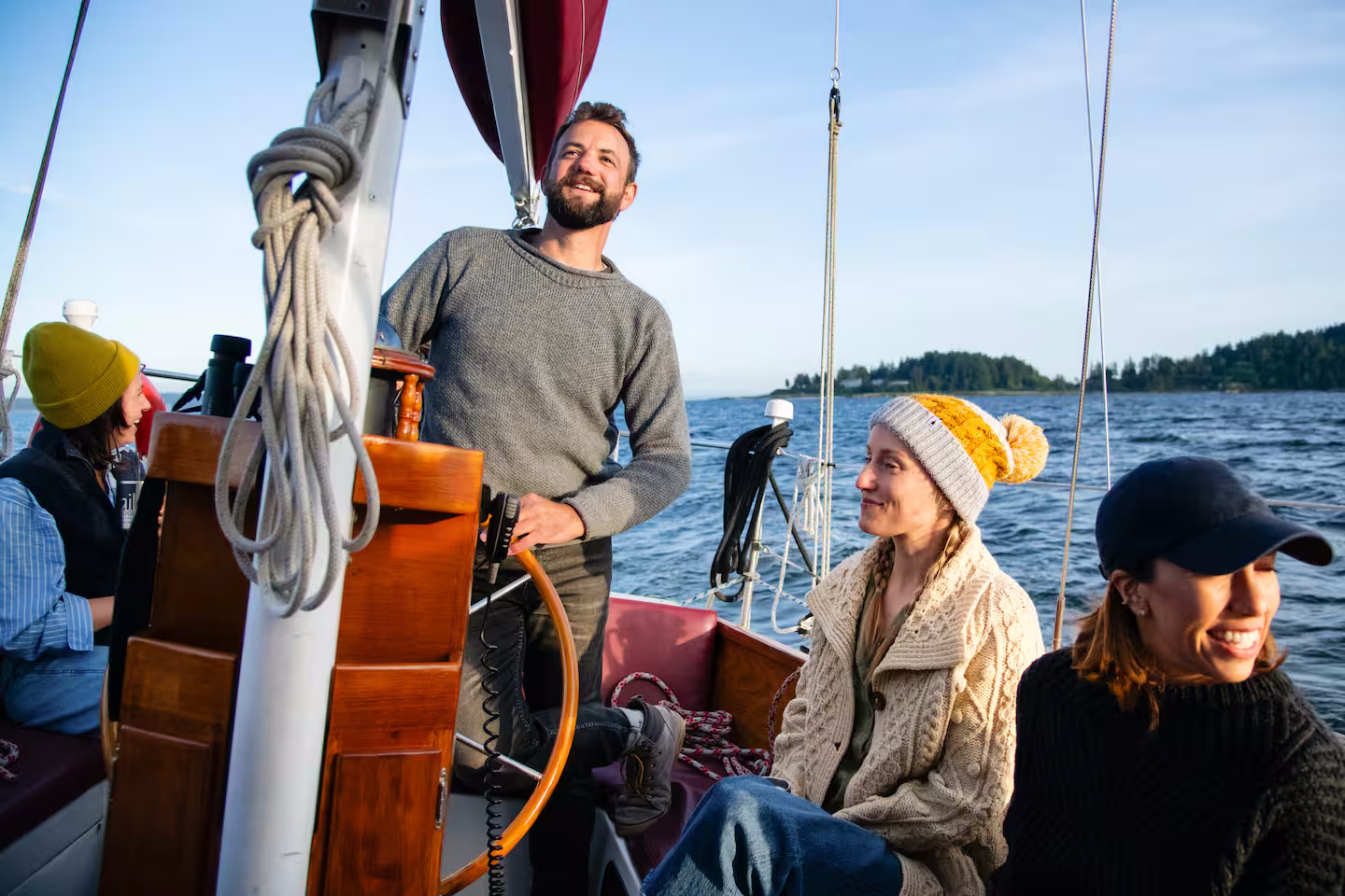 Smiling man at the helm steering a sailboat with two women seated nearby, blue ocean and distant island in the background.