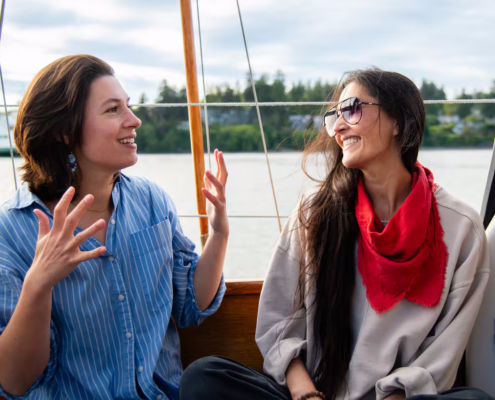 Two women sit on a boat, smiling and talking; one in a blue striped shirt raises hands, the other wears sunglasses and a red scarf.