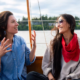 Two women sit on a boat, smiling and talking; one in a blue striped shirt raises hands, the other wears sunglasses and a red scarf.