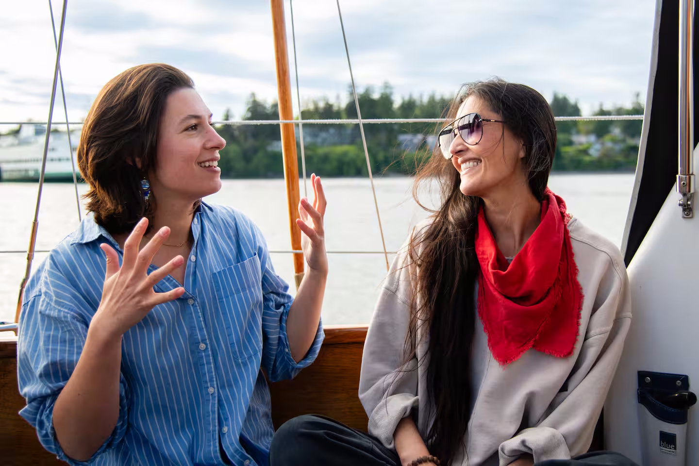 Two women sit on a boat, smiling and talking; one in a blue striped shirt raises hands, the other wears sunglasses and a red scarf.