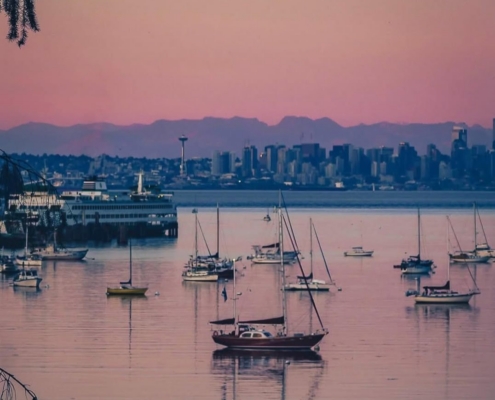 Sailboats gently anchored in a calm harbor at sunset with a pink-orange sky and a distant city skyline.