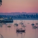 Sailboats gently anchored in a calm harbor at sunset with a pink-orange sky and a distant city skyline.