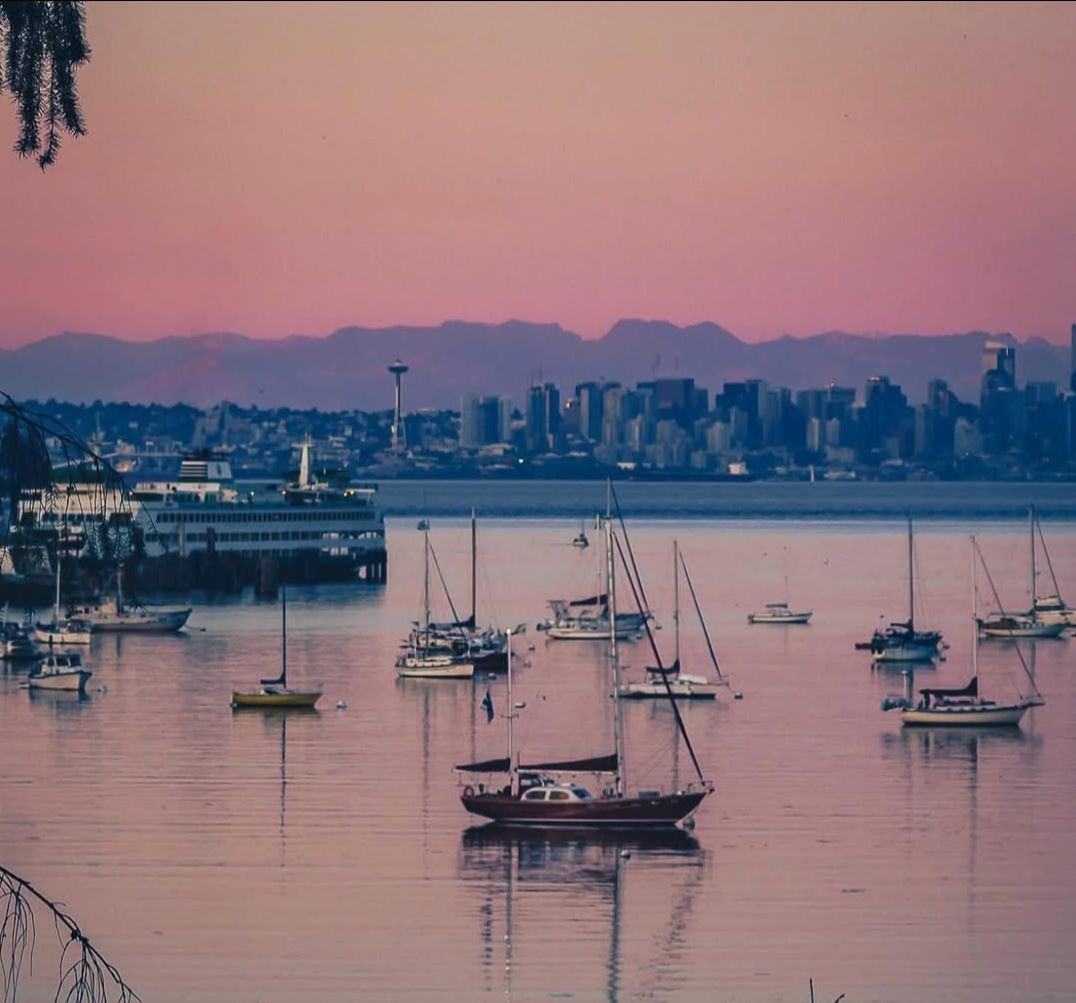 d6cbe48c-918e-91ec-5207-99b92a42ee32 | Sail Bainbridge | Captained Sailing Adventures | Bainbridge Island | Seattle Sailboats gently anchored in a calm harbor at sunset with a pink-orange sky and a distant city skyline.