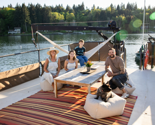 Family of four with a dog relaxing on a boat deck around a wooden table with flowers by a lake.