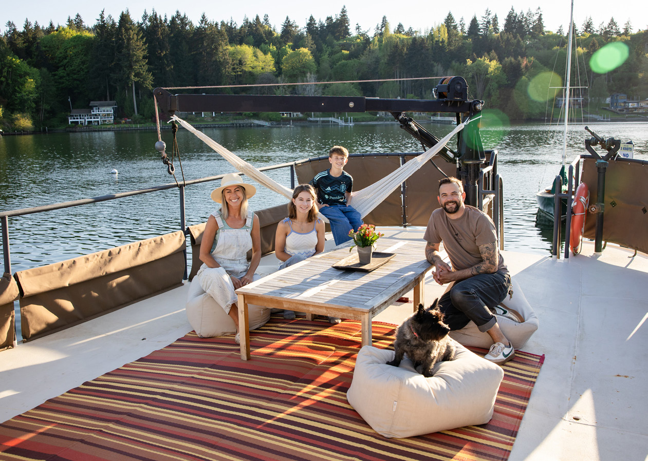 Family of four with a dog relaxing on a boat deck around a wooden table with flowers by a lake.