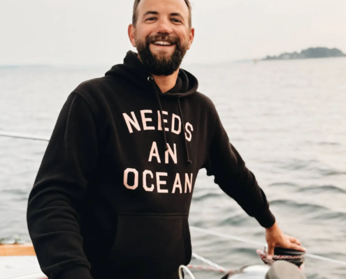 Smiling man in a black hoodie at the helm of a boat, gripping a wooden steering wheel with open ocean behind him.