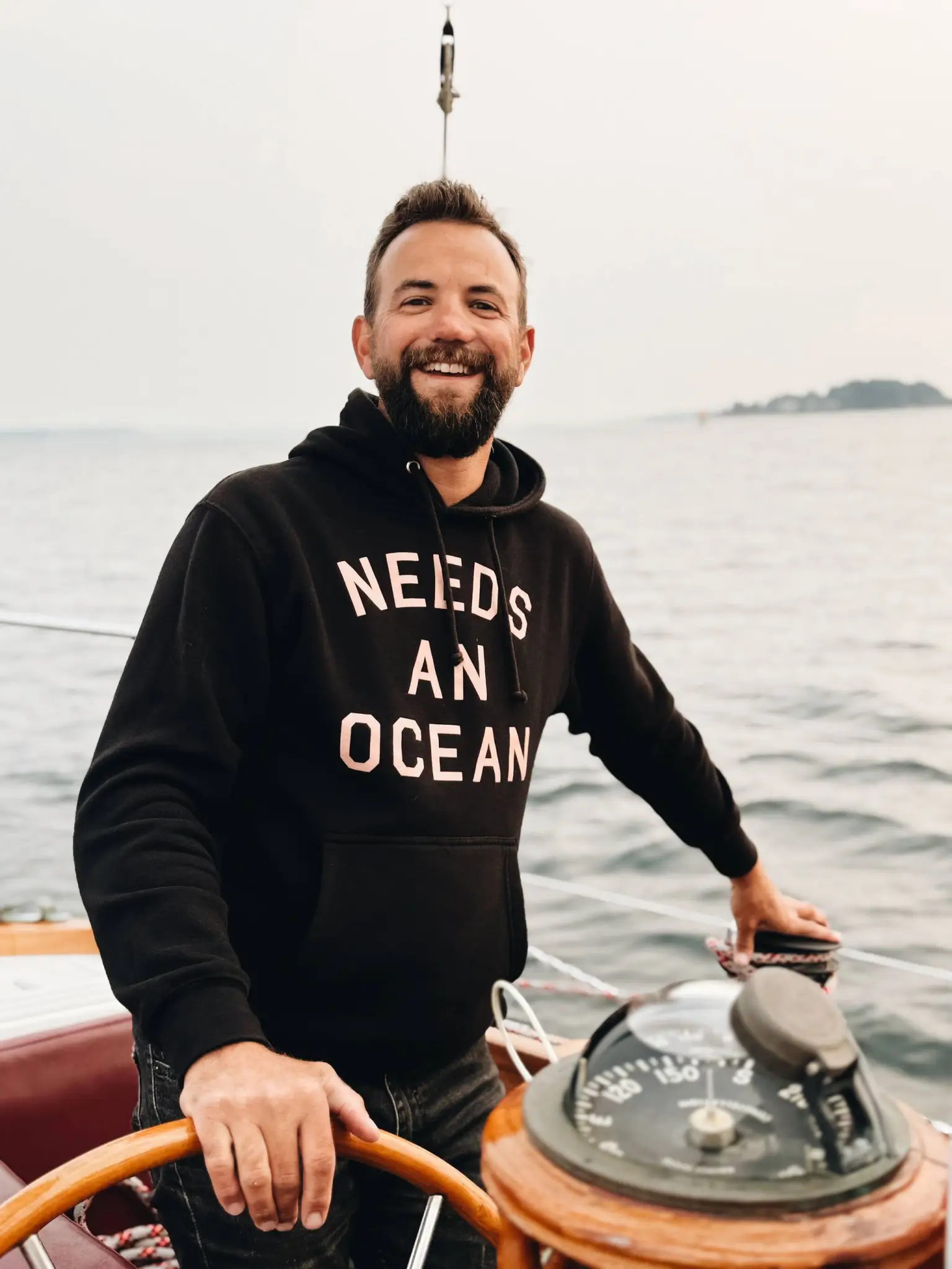 Smiling man in a black hoodie at the helm of a boat, gripping a wooden steering wheel with open ocean behind him.
