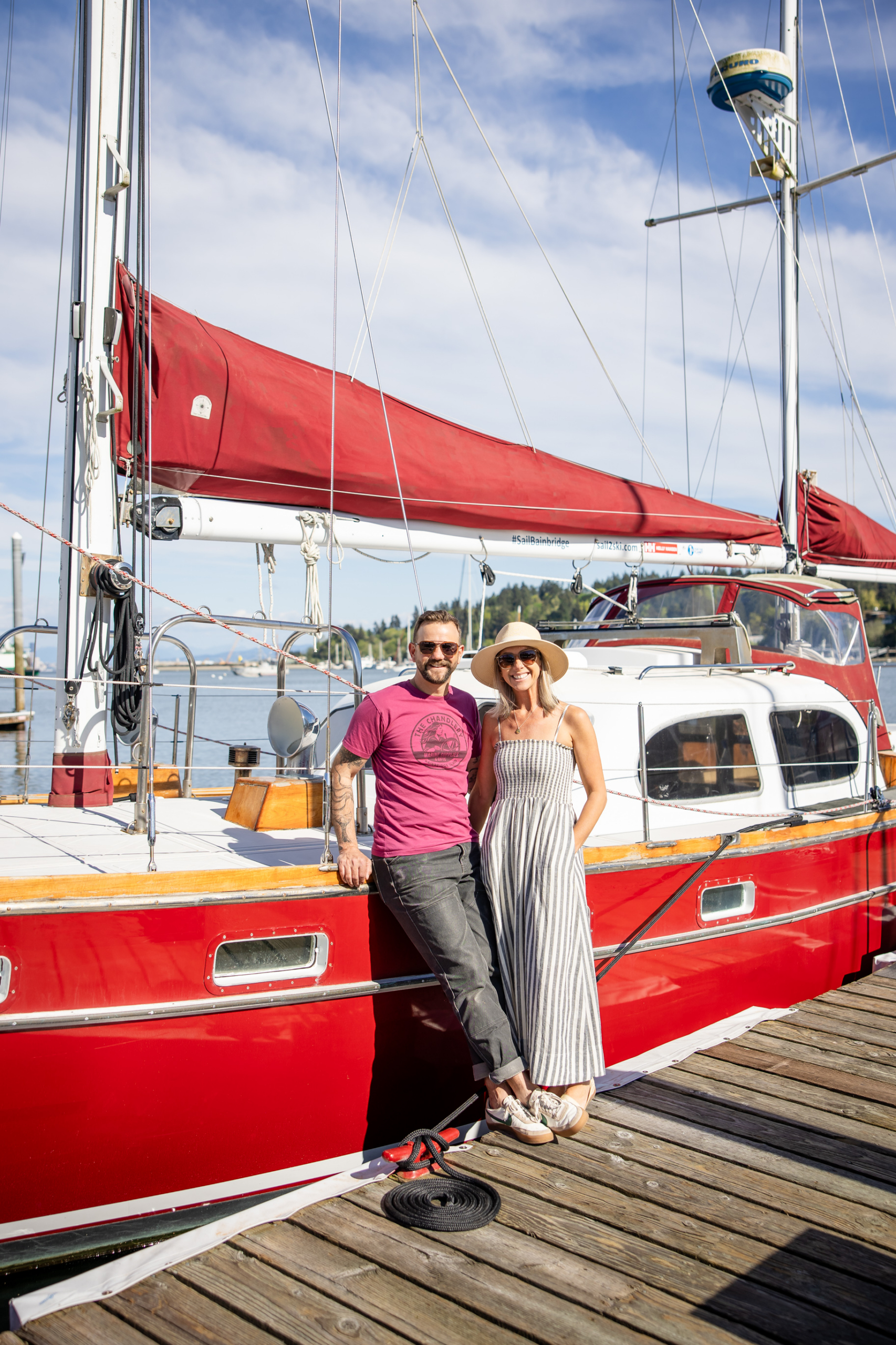 6a29b3ced1efb11e7cad0a63_202604Chandlery-114-1 | Sail Bainbridge | Captained Sailing Adventures | Bainbridge Island | Seattle A couple stands on the deck of a red sailboat at a marina, smiling with rigging and blue sky in the background.