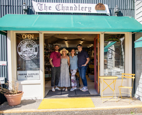 Four people stand in the open doorway of a marine-supplies shop named The Chandlery, with a green awning and a yellow table outside.