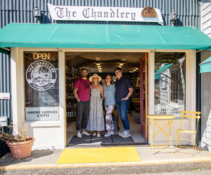 6a29b52953ba2270ee7005dc_chand 2 | Sail Bainbridge | Captained Sailing Adventures | Bainbridge Island | Seattle Four people stand in the open doorway of a marine-supplies shop named The Chandlery, with a green awning and a yellow table outside.